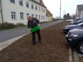 Mann mit Wanne streut Saatgut auf erdige Fläche neben Parkplatz