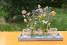 5 Gläser auf einem Tablett, in zwei davon ist ein Blumenstrauß 